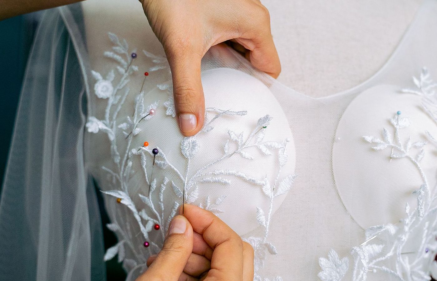 Licensed stock photograph: two hands setting pins into the lace bodice of a wedding gown on a dress form, coloured pin heads along the seam.